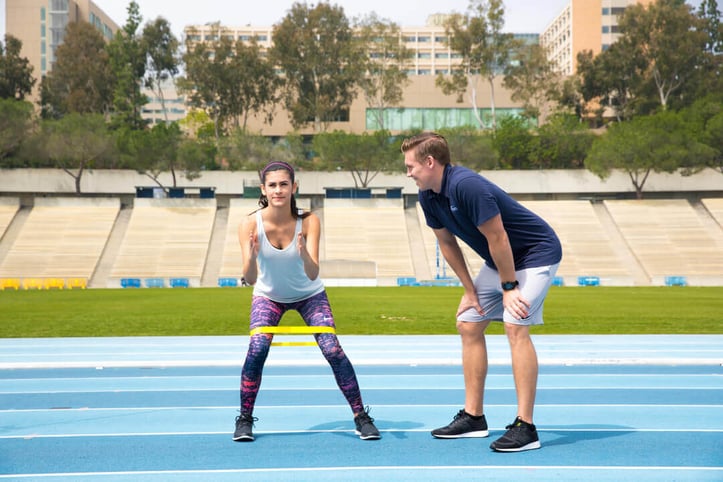 Male NASM trainer assisting female client with band workout outside